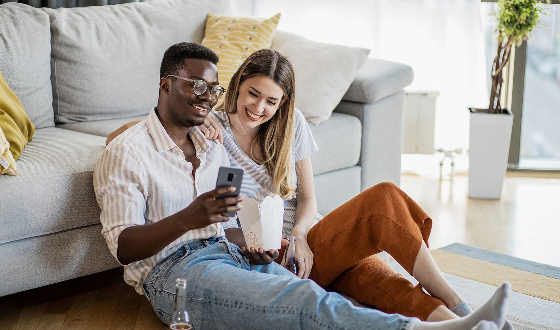 a man and woman with take out, sit against a couch on their living room floor and watch at their cell phone